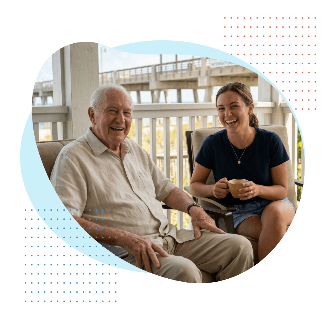 A senior man and a cheerful companion laughing together on a shaded porch in Deerfield Beach, with the International Fishing Pier visible in the background.